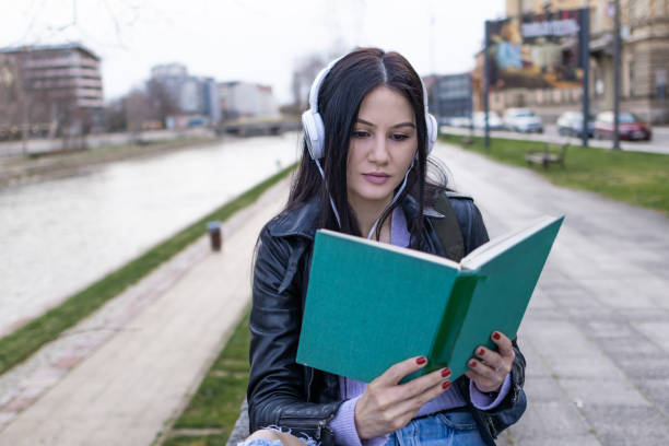 istock Young beautiful female university student with backpack is enjoying near the river. She is wearing headphones and leather jacket while waiting for her college class to start. 2132876344
