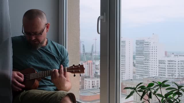 A bearded guy plays music with inspiration, enjoys his hobby. Adult caucasian man playing the ukulele sitting on the windowsill in a city apartment.
