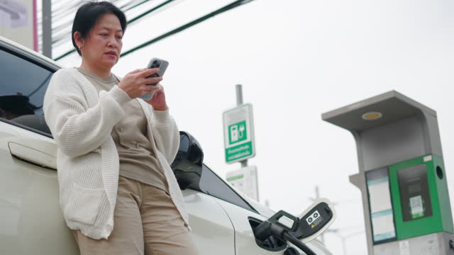 Asian woman use mobile phone connect to see status and waiting when charge  ev car at electronic charging station