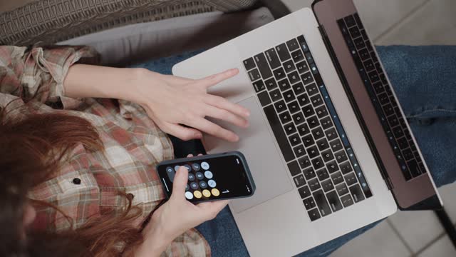 Above view of a young woman's hands calculating a budget on her phone and laptop