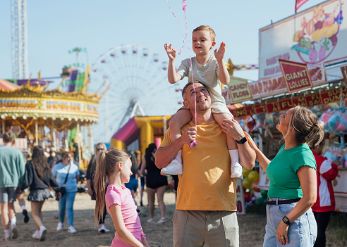 Family visiting an outdoor travelling carnival with different rides and food stalls in the North East of England. The dad is holding his son on his shoulders.