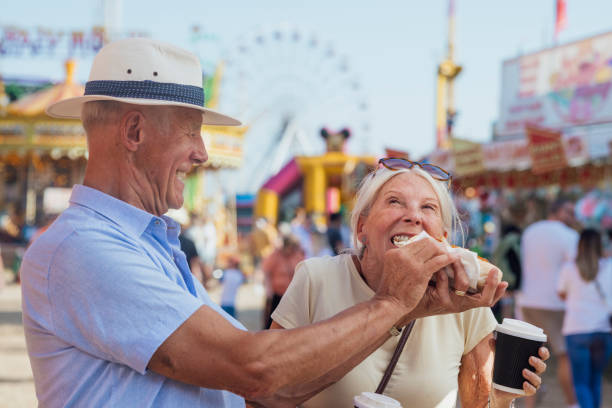 Senior couple eating a hot dog together while at an outdoor travelling carnival in the North East of England. They are enjoying their lunch in the sunshine and the woman is holding a takeaway hot drink.