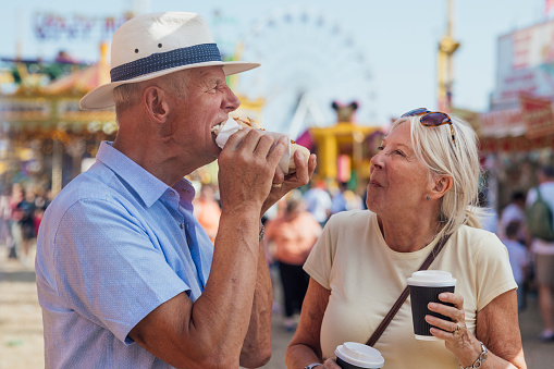 Senior couple eating a hot dog together while at an outdoor travelling carnival in the North East of England. They are enjoying their lunch in the sunshine and the woman is holding a takeaway hot drink.
