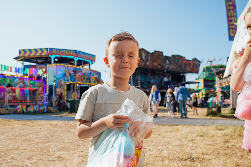 Young boy eating candy floss while his family visit an outdoor travelling carnival with different rides and food stalls in the North East of England. He is standing in the sun with his eyes closed.