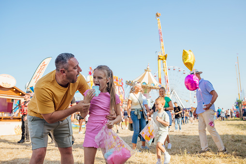 Family visiting an outdoor travelling carnival with different rides and food stalls in the North East of England. A father and daughter are sharing candy floss while the rest of the family are behind them.