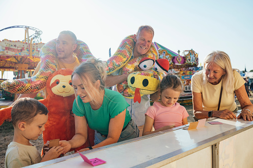 Multi generation family visiting an outdoor travelling carnival with different rides and food stalls in the North East of England. They are playing a carnival game together, trying to win a prize. Some members of the family are holding big stuffed toys they have already won.