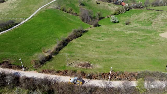 Yellow single smooth drum ride-on vibratory roller compactor moves along a rocky road through the village where it builds infrastructure, meadows and electric poles in the background, drone shot
