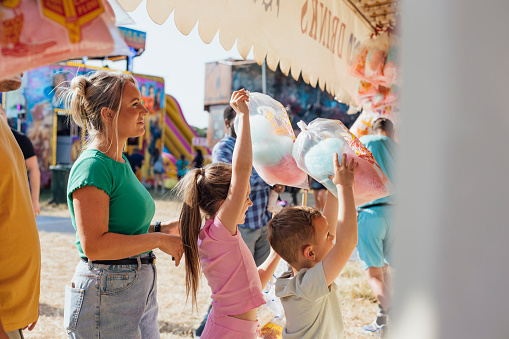 Family visiting an outdoor travelling carnival with different rides and food stalls in the North East of England. They are visiting a van selling confectionary. The children are holding bags of candy floss in the air.