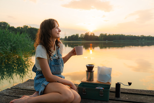 Woman  sitting near the lake on wooden pier   and brewing coffee at sunset
