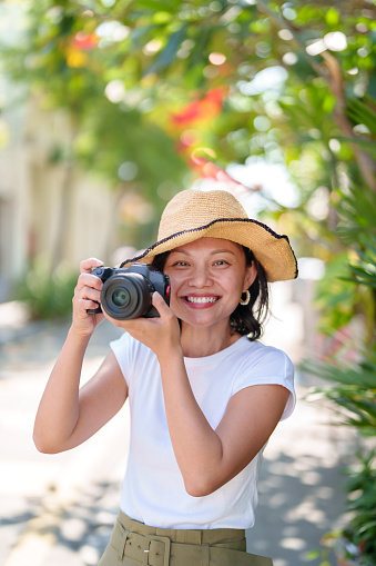 A solo Asian woman traveler, wearing a straw hat, takes photos with her camera while exploring Georgetown, Penang. She travels light, capturing the lively streets and colorful buildings. It's a simple, joyful way for her to explore and make memories.