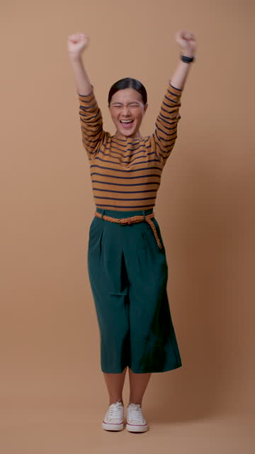 Asian woman happy confident showing her fist make a winning gesture standing isolated over beige background.