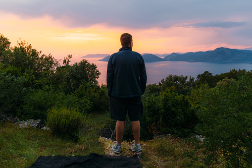 Rear view of a male contemplating view from above of Mediterranean sea in pink orange sunset lights with the hills of Oludeniz in Turkey