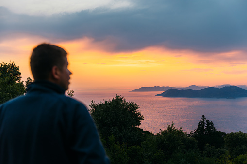 Rear view of a male contemplating view from above of Mediterranean sea in pink orange sunset lights with the hills of Oludeniz in Turkey