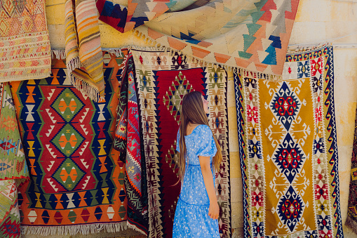 Side view of a happy female with long hair and in a blue dress walking by the colourful authentic Turkish carpets in the background in Göreme, Middle East