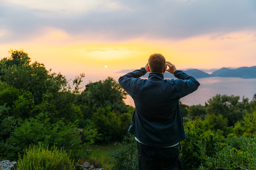 Rear view of a male photographing the Mediterranean sea with the hills of Oludeniz during dramatic colorful sunset in Mugla province, Turkey