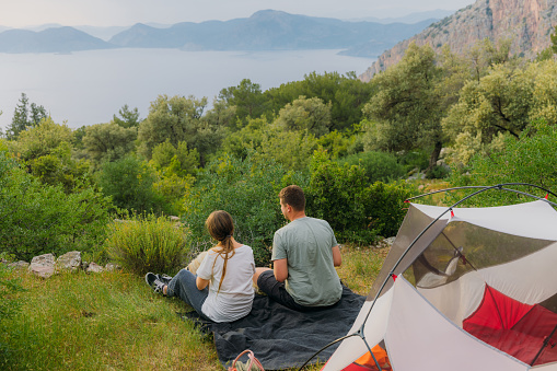 Rear view of heterosexual couple with their dog camping with a red tent in the green forest with a view of the Mediterranean sea and the mountains during scenic sunset in Oludeniz, Turkey
