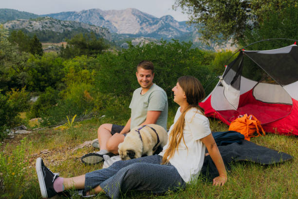 Side view of heterosexual couple with their dog camping with a red tent in the green forest with a view of the Mediterranean sea and the mountains during scenic sunset in Oludeniz, Turkey
