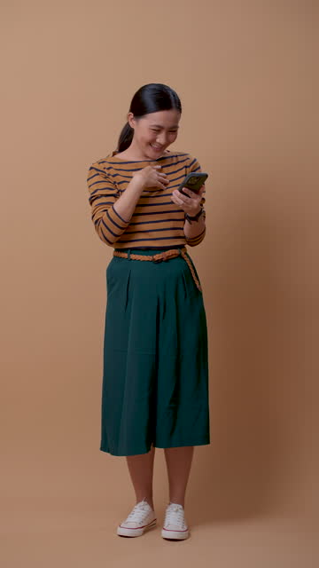 Asian woman laughing enjoyed using smartphone isolated over beige background.