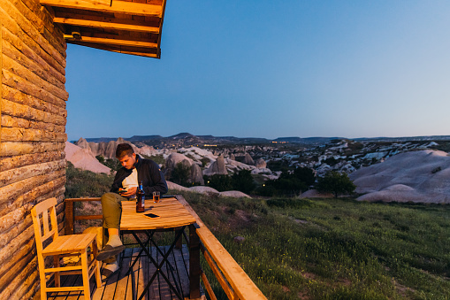Male enjoying drinks at the terrace using phone for work with scenic view of the rock formations on the hills during blue hour in Capapdocia, Turkey