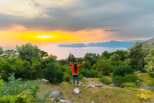 Happy female raising arms with the viewpoint of the Mediterranean sea with the hills of Oludeniz during dramatic colourful sunset in Mugla province, Turkey
