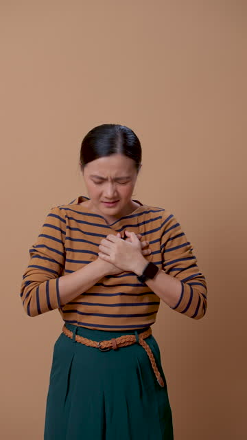 Asian woman suffering from chest pain standing isolated on beige background.