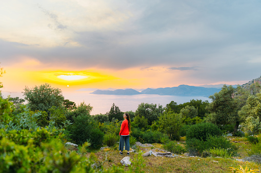 Happy female in orange contemplating viewpoint of the Mediterranean sea with the hills of Oludeniz during dramatic colourful sunset in Mugla province, Turkey