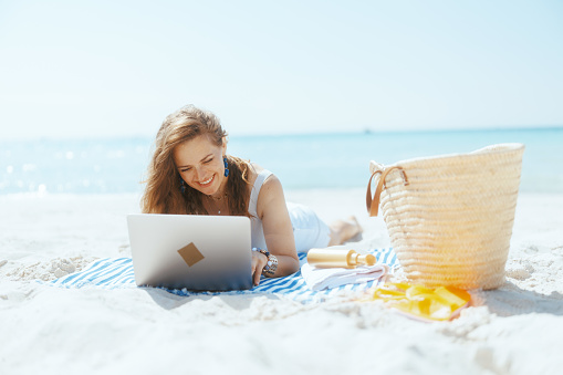 smiling modern woman on the ocean coast with straw bag and striped towel using laptop.