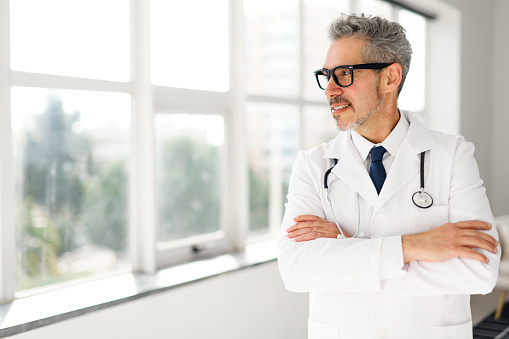 Senior doctor in white coat and stethoscope, suggest a comfortable and patient-centric clinic environment. The soft, natural lighting contributes to a calm and inviting atmosphere for consultation