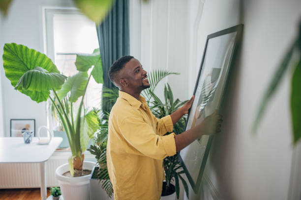 Smiling man adjusting picture frame at home Cheerful black man decorating his cozy living space with artwork hanging art on wall stock pictures, royalty-free photos & images
