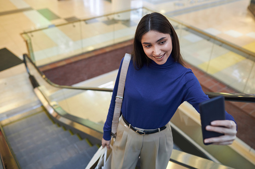 High angle portrait of smiling young woman taking selfie photo or filming video standing on escalator in shopping mall and holding bags copy space