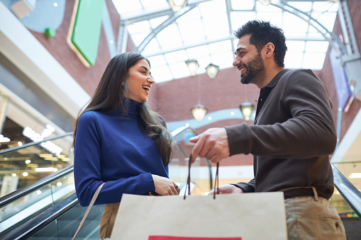 Waist up portrait of smiling young couple standing on escalator in shopping mall and holding bags while enjoying shopping spree together
