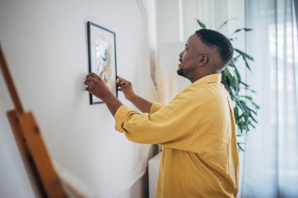 Man hanging artwork at home Focused black man carefully places a framed picture on his living room wall hanging art on wall stock pictures, royalty-free photos & images