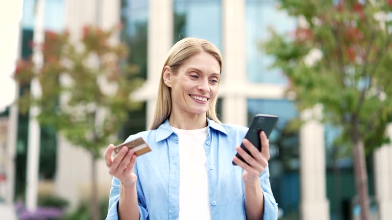 Smiling female is shopping online using credit card and phone standing on the city street near a modern building.