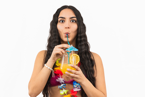 young Latin woman wearing a bikini looks at the camera with wide eyes as she drinks a summer cocktail with a straw, radiating excitement and enjoyment on white background