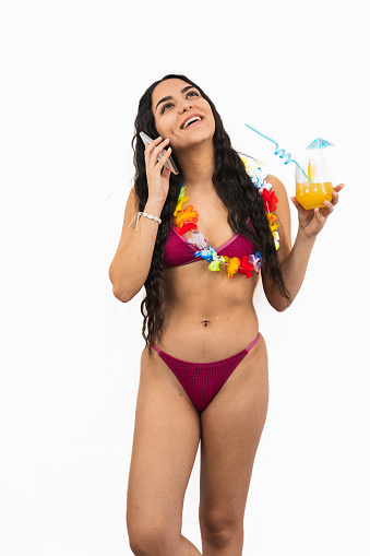 vertical portrait brunette Latin girl wearing a bikini is talking on her cellphone while holding an orange soda. She appears relaxed and engaged in conversation in the beach on white background