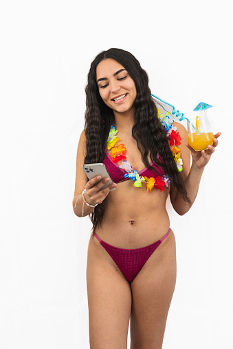 vertical portrait Latin girl is seen texting on her smartphone while wearing a bikini and a flower necklace. She appears relaxed and immersed in her phone on white background