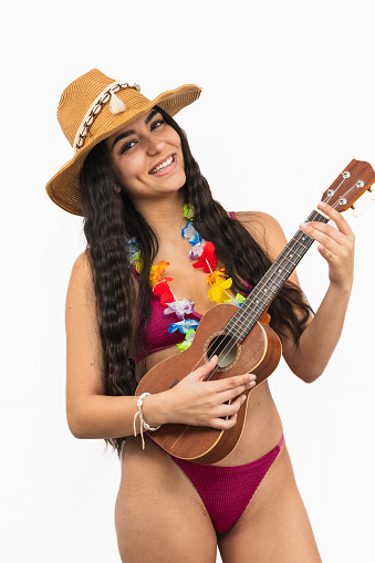 vertical portrait closeup smiling Latina woman in a bikini is seen playing the ukulele. She is wearing a flower necklace and a beach hat, radiating joy on a sunny beach day on white background