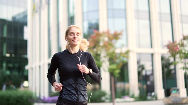 Adult female runner with headphones jogging in city street. Fit mature sportswoman in a black tracksuit enjoys jogging outdoors on park alley while listening to music
