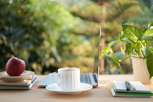 A cup of tea on a wooden work table outside with side view of Laptop and notebooks balcony outdoor