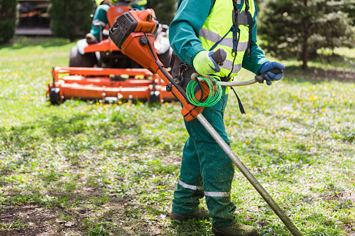 A professional landscaper cuts grass with a string trimmer, wearing safety gear with a ride-on mower in the background