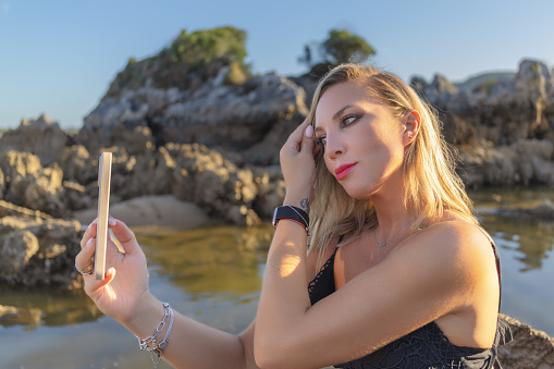 An attractive woman enjoys on the beach in the rays of the setting sun and takes a selfie