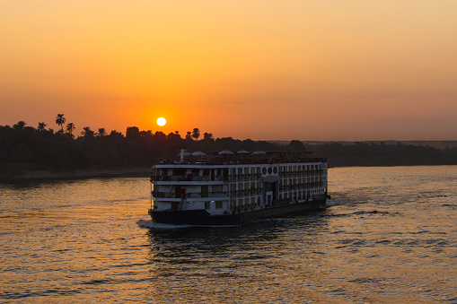 Sunset on the Nile River with a cruise ship sailing on the water and a grove of trees behind