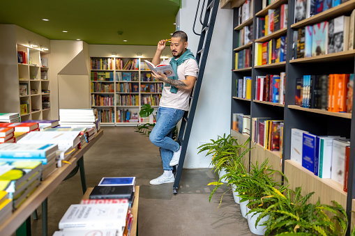 Reading a book. Young asian man standing near the book shelves and reading a book