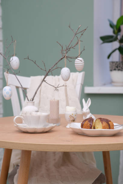 istock Easter table. Vase with fresh branches, cup of tea, cupcake on plate, boiled egg, ceramic bunny, candle on wooden table with white chair on curtain background. 2133387528