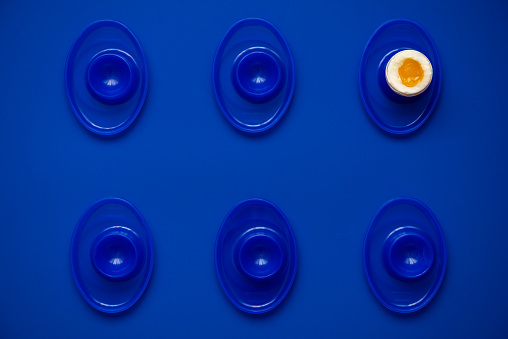 Monochromatic flat lay of evenly spaced blue egg cups with a single soft boiled egg on a blue background