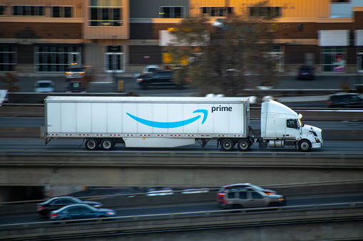 An Amazon Prime independent contractor driving a semi truck on the freeway through an urban city center. The truck is owned by the contractor, and the trailer is owned by Amazon.