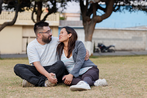 Hispanic couple sitting in a field looking at each other - young couple in love in the park - newly married couple - Latin American adult couple