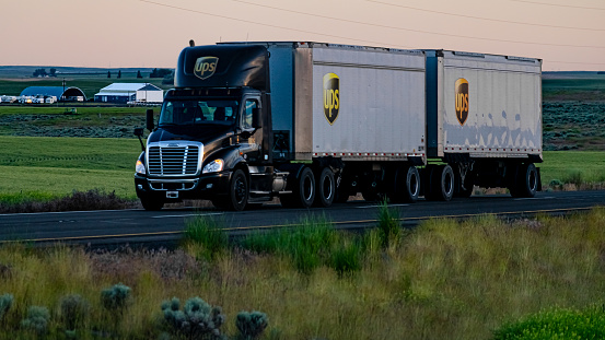 UPS feeder semi-truck with double trailers, on the freeway passing by farm fields.