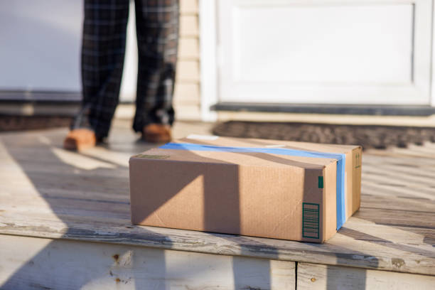 Brown cardboard parcel box with address label lying on wooden porch at home entrance. Unrecognizable woman's legs in trousers seen in background approaching to pick up delivered package