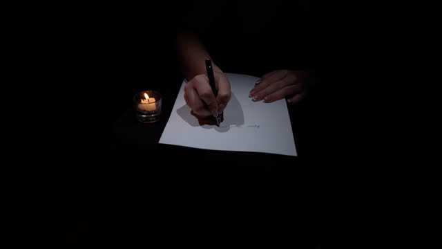 Hand Writes with a Pen on White Paper in a Dark Room by Candlelight